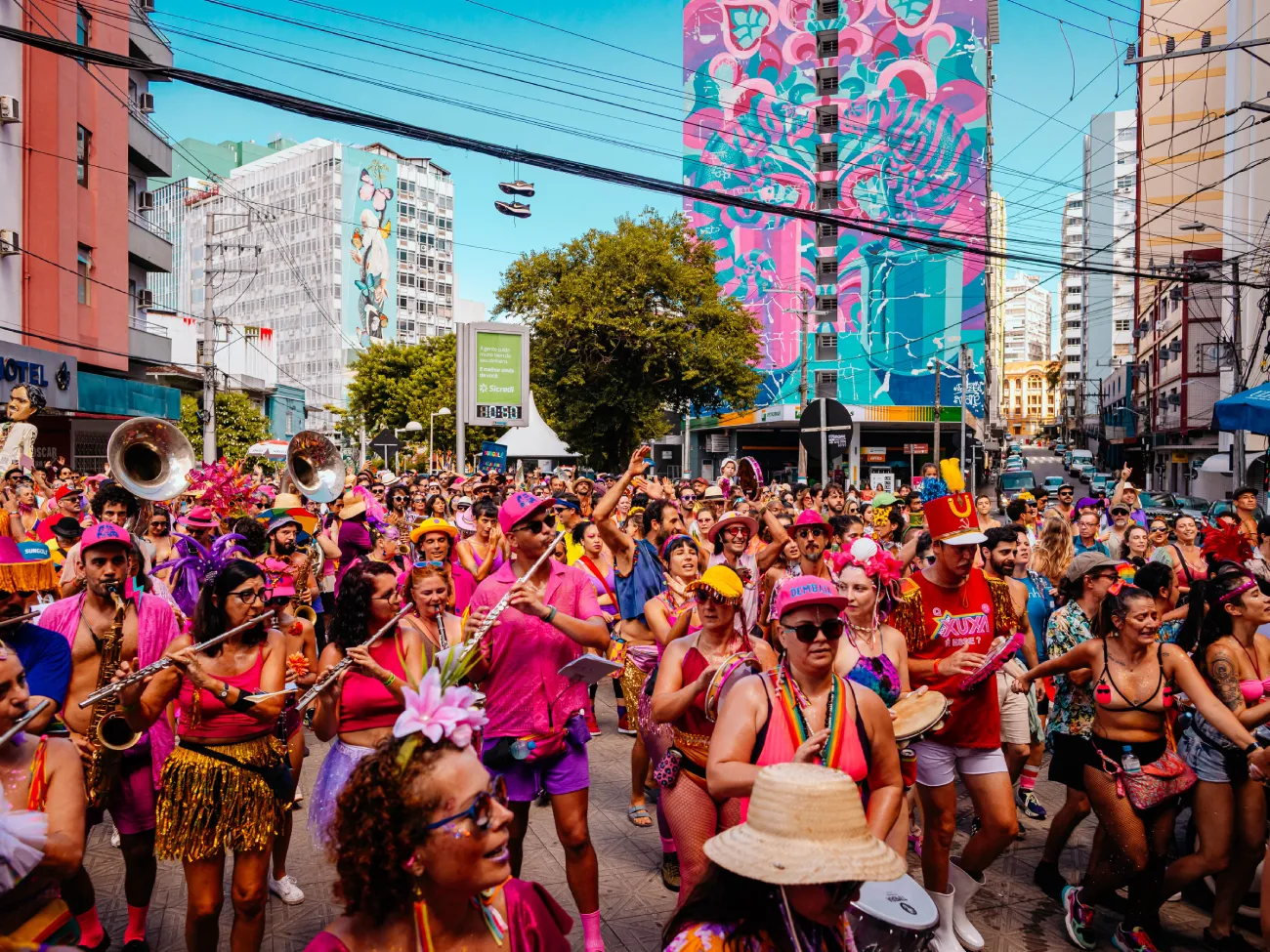A Fanfarra da Ponte abre a programação pré-Carnaval em Florianópolis neste domingo, 1º de fevereiro, com um cortejo que parte da cabeceira da Ponte Hercílio Luz, no lado continental, e segue pela Avenida Beira-Mar Continental. A concentração está marcada para as 10h30, e a saída oficial ocorre ao meio-dia, integrando o Festival Ponte Que Pariu. O evento marca o início das atividades públicas do bloco em 2026 e propõe ocupar um dos principais cartões-postais da cidade com música, fantasias e participação popular, reunindo moradores e visitantes em um desfile a céu aberto. FANFARRA DA PONTE ABRE O PRÉ-CARNAVAL NA PONTE HERCÍLIO LUZ A escolha da Ponte Hercílio Luz como ponto de partida reforça o caráter simbólico da apresentação. Considerada um dos principais marcos urbanos da capital catarinense, a estrutura passa a ser também cenário de um encontro cultural que une tradição carnavalesca e uso do espaço público. O cortejo percorre a Beira-Mar Continental, com instrumentos de sopro e percussão, promovendo interação direta com o público ao longo do trajeto. A proposta do bloco é valorizar o Carnaval de rua como manifestação democrática, acessível e integrada ao cotidiano da cidade. MÚSICA, COLETIVIDADE E OCUPAÇÃO DO ESPAÇO PÚBLICO De acordo com o regente e professor da Fanfarra da Ponte, Ricardo Lagartixa, a abertura do pré-Carnaval nesse local tem significado especial para o grupo. “Abrir o pré-Carnaval na Ponte Hercílio Luz tem um significado muito forte pra gente. A ponte é encontro, é travessia, é símbolo da cidade, e o Bloco da Fanfarra, nasceu exatamente com essa ideia de ocupar a rua, aproximar as pessoas e celebrar Florianópolis com alegria e respeito. Somos cerca de 80 integrantes no bloco e, em alguns momentos, ainda contamos com a força dos alunos da EPP, o que transforma o cortejo num grande coletivo em movimento. É música, é troca, é Carnaval acontecendo de forma viva, popular e acessível”. Segundo a organização, o desfile deve reunir integrantes do bloco e estudantes vinculados às atividades formativas da fanfarra, ampliando o caráter educativo e coletivo da apresentação. EVENTO INTEGRA O FESTIVAL PONTE QUE PARIU A saída da Fanfarra da Ponte faz parte da programação do Festival Ponte Que Pariu, projeto incentivado pelo Brasil Atacadista e pela Brahma, com apoio do Programa de Incentivo à Cultura da Fundação Catarinense de Cultura (FCC) e do Governo do Estado de Santa Catarina. A iniciativa marca o início do calendário oficial de apresentações do bloco em 2026, que tradicionalmente atrai milhares de foliões ao longo do verão, consolidando-se como uma das manifestações culturais ligadas ao Carnaval de rua em Florianópolis. HISTÓRIA DA FANFARRA DA PONTE A Fanfarra da Ponte surgiu no Carnaval de 2020, quando um pequeno grupo de músicos decidiu levar seu estandarte e seus instrumentos pelas ruas do Centro da capital catarinense. A partir dessa experiência, o coletivo se estruturou e ampliou suas atividades. Em 2022, foi criado o Núcleo de Estudos Fanfarra da Ponte, voltado ao ensino gratuito de instrumentos de sopro e percussão. Desde então, integrantes da fanfarra e alunos do núcleo participam conjuntamente dos desfiles, com repertório próprio baseado na música brasileira. O grupo se apresentou nos Carnavais de 2023, 2024 e 2025, fortalecendo sua presença na cena cultural da cidade e consolidando o formato de cortejo musical como parte da programação pré-carnavalesca. SERVIÇO O quê: Cortejo pré-Carnaval da Fanfarra da Ponte – Festival Ponte Que Pariu Quando: Domingo, 1º de fevereiro de 2026 Onde: Concentração na cabeceira da Ponte Hercílio Luz (lado continental) e percurso pela Beira-Mar Continental Ingressos: Evento gratuito, sem necessidade de retirada prévia Mais informações em: redes sociais oficiais da Fanfarra da Ponte e do Festival Ponte Que Pariu