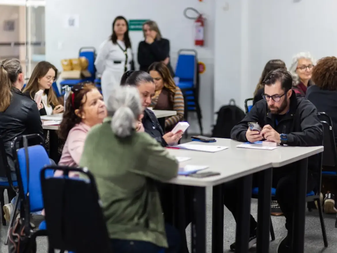 A imagem mostra um grupo de pessoas sentadas em mesas e cadeiras em uma sala de reunião ou auditório participando do projeto Desbrave Chapecó